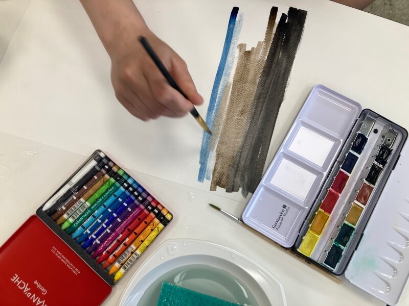 Eine Person malt mit Aquarellfarben und Wachskreiden auf einem Blatt Papier, ruhige Farbtöne und konzentrierte Handbewegung deuten auf kreativen Ausdruck hin.