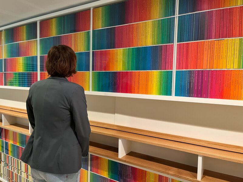Eine Person betrachtet ein großformatiges, farblich sortiertes Wandregal mit Buchrücken in Regenbogenfarben in der Siegfried-Unseld-Bibliothek.